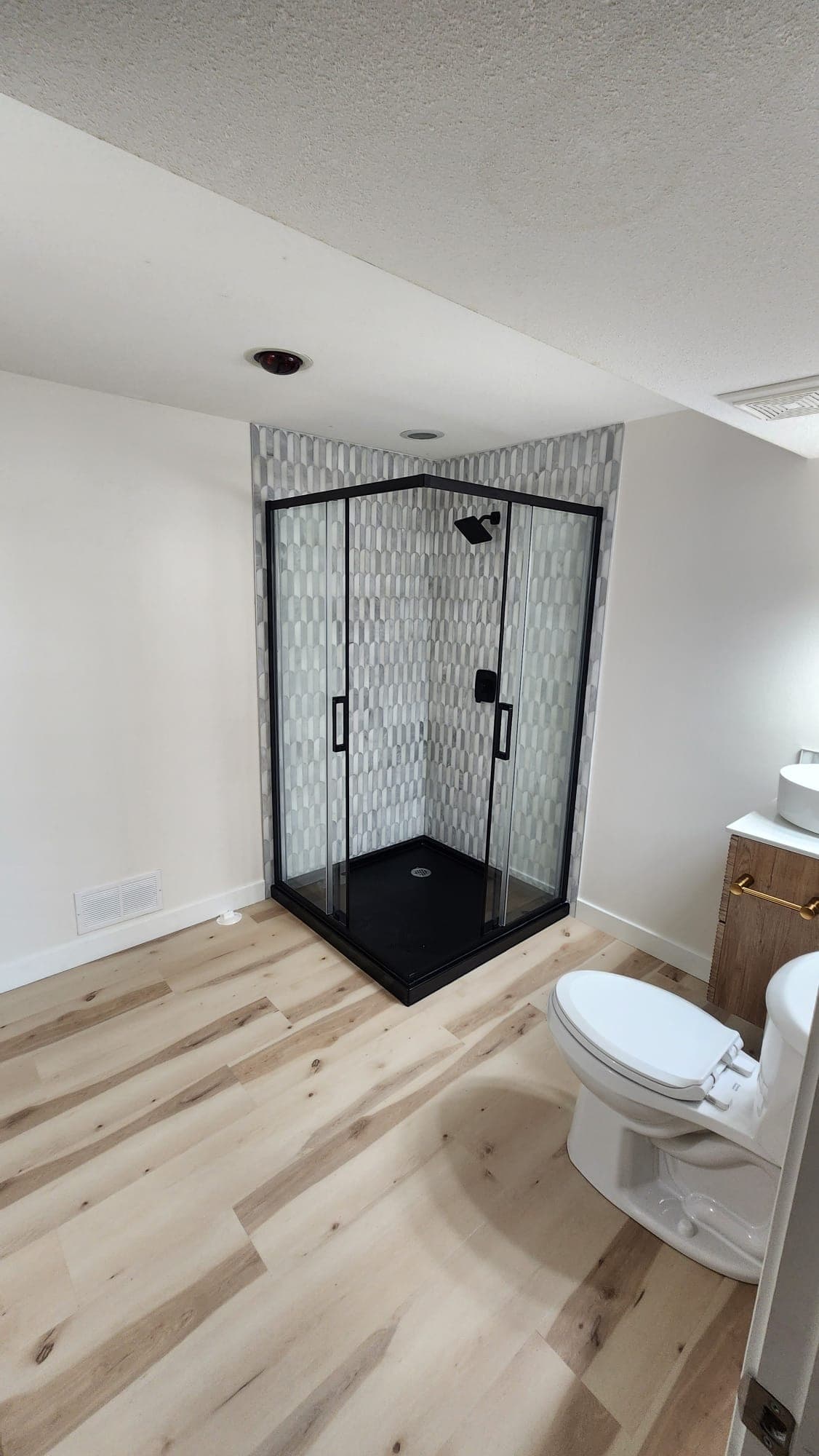 Bright basement bathroom with white walls, black glass shower and vinyl plank floor, Calgary